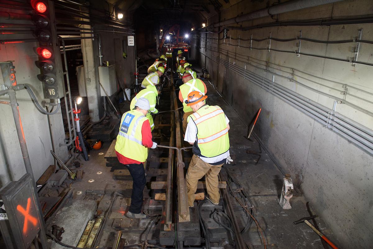 Nearly a dozen crew in safety gear all standing on each side of rail track. They are holding the track.