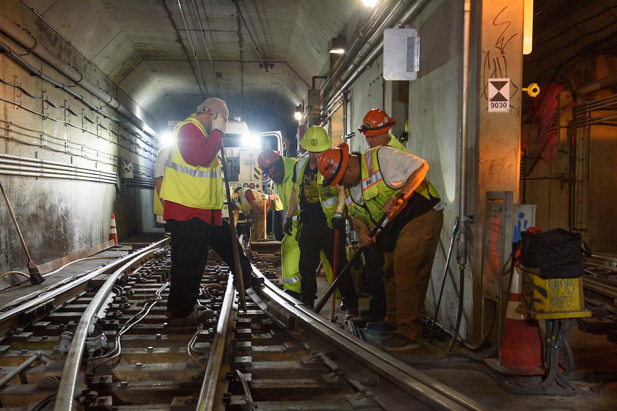 Several crew in safety gear standing over a rail track with tools angled in hand.