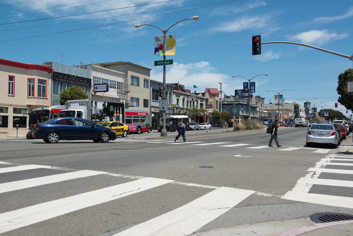 Photograph of Geary Boulevard at 4th Avenue where two pedestrians are crossing in the crosswalk while a car makes a left turn.