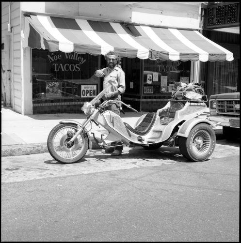 Vintage black and white photo of a person standing over a motorcycle in front of a restaurant "Noe Valley Tacos"