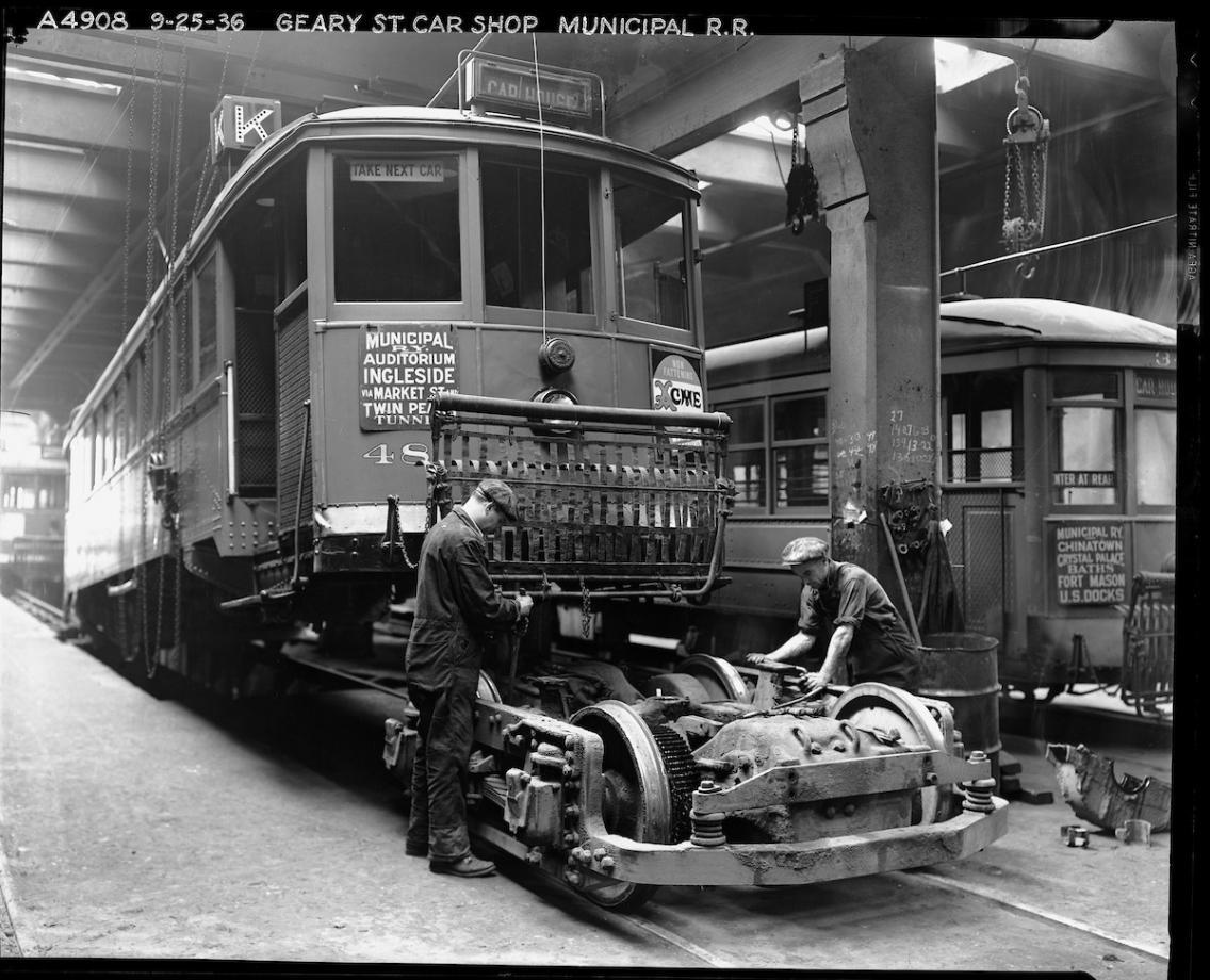 Inside the shops at Geary Car House, two mechanics work on a truck (the combined wheels, suspension, and motor of the streetcar)