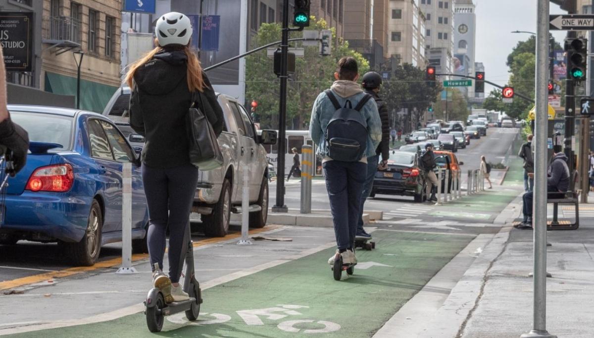 photo of 2 individuals riding powered scooters, one riding a skateboard, and pedestrians