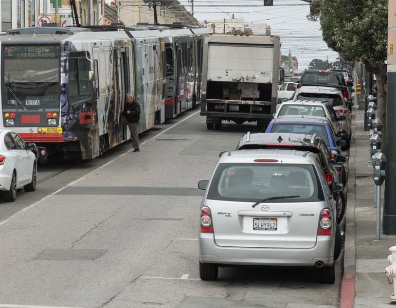 Parked cars sit along the curb on Taraval Street as a customer boards a Muni L Taraval train in the roadway.