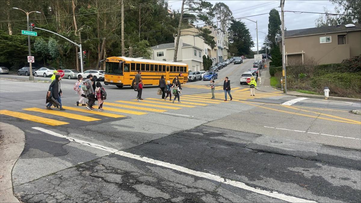 students using a crosswalk to cross clarendon avenue at panorama drive a bus is waiting in the intersection.