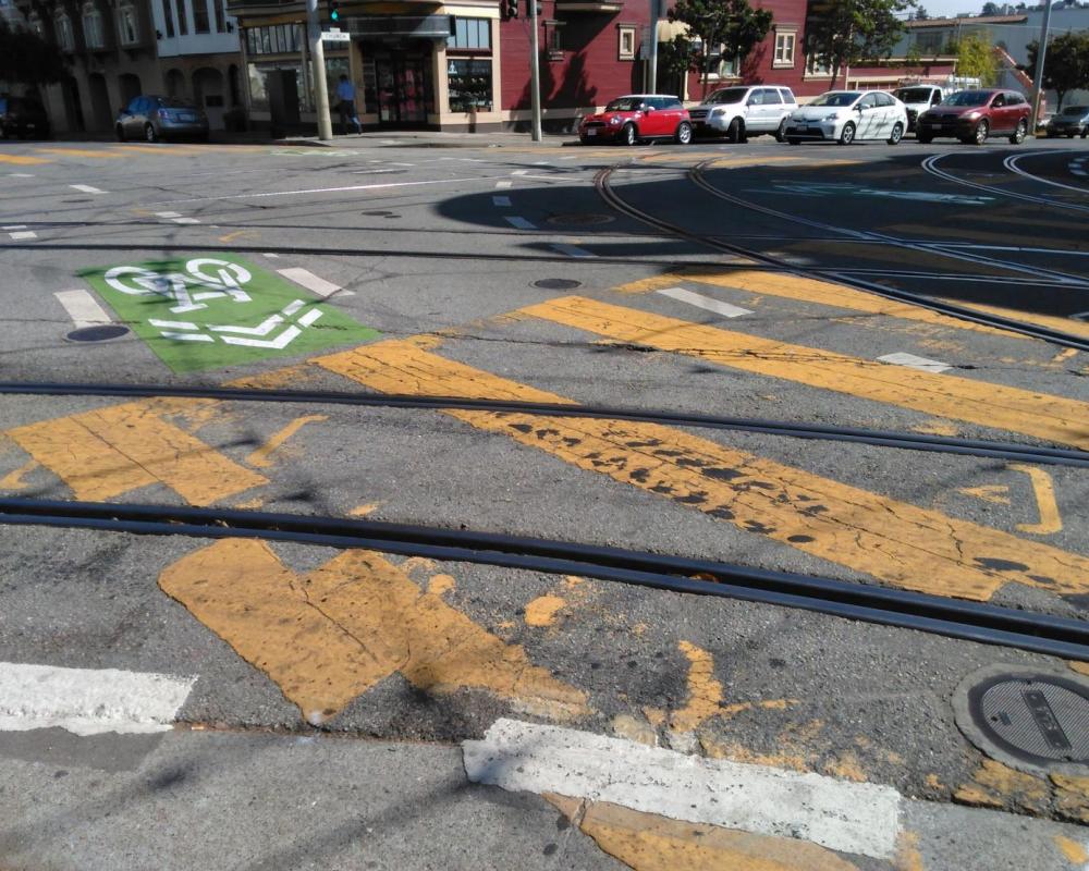 Green-backed sharrows, yellow continental crosswalks, and other roadway markings are overlaid with tracks in the roadway