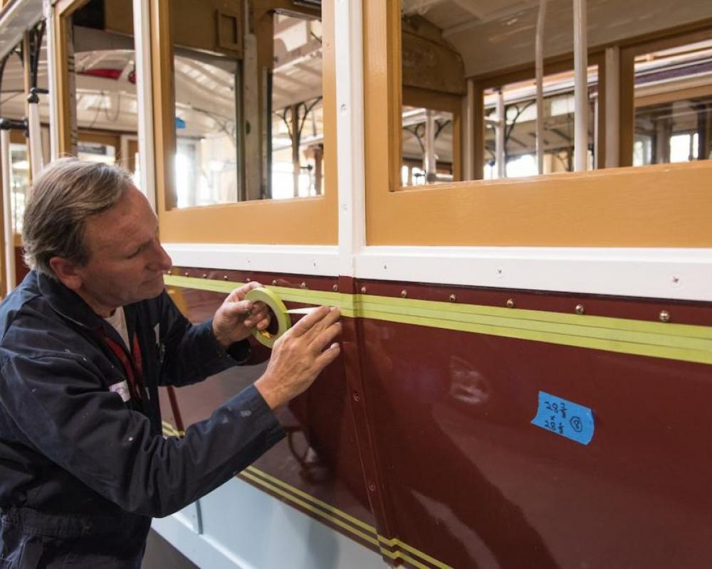 A painter lays down masking tape to paint striping on the side of Powell St. Cable Car 22,