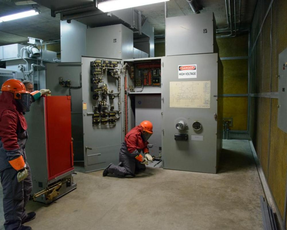 Two crew members in full protected wear inside a shops location. The walls are bordered with systems cabinets and decks. 