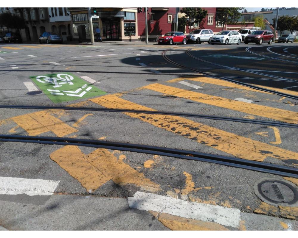Green-backed sharrows, yellow continental crosswalks, and other roadway markings are overlaid with tracks in the roadway