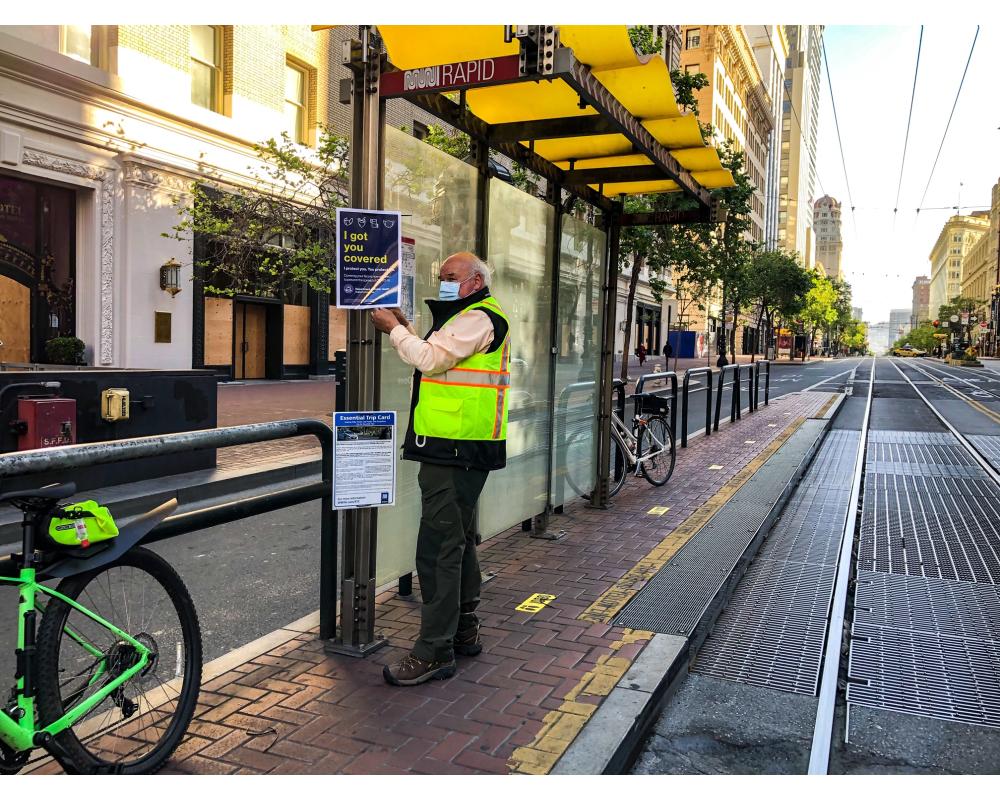 Photo of a Market Street Ambassador posting new face mask, ETC, and wayfinding signage on a boarding island bus stop.