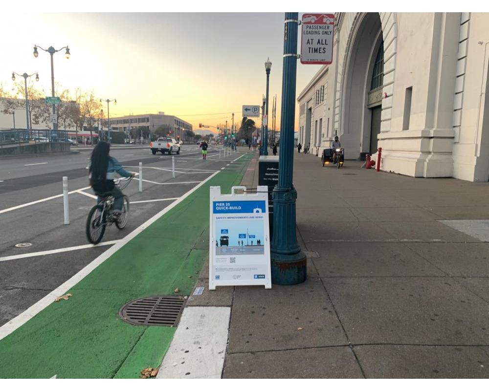 person riding bike on green bike path past Ferry Building and Quick Build sandwitch board