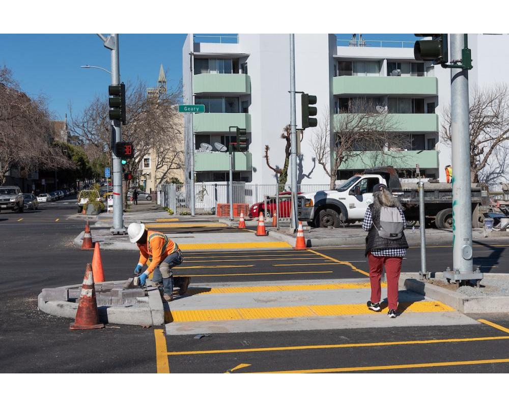 A worker installs decorative pavers at a new median refuge while a woman crosses Geary and Steiner.