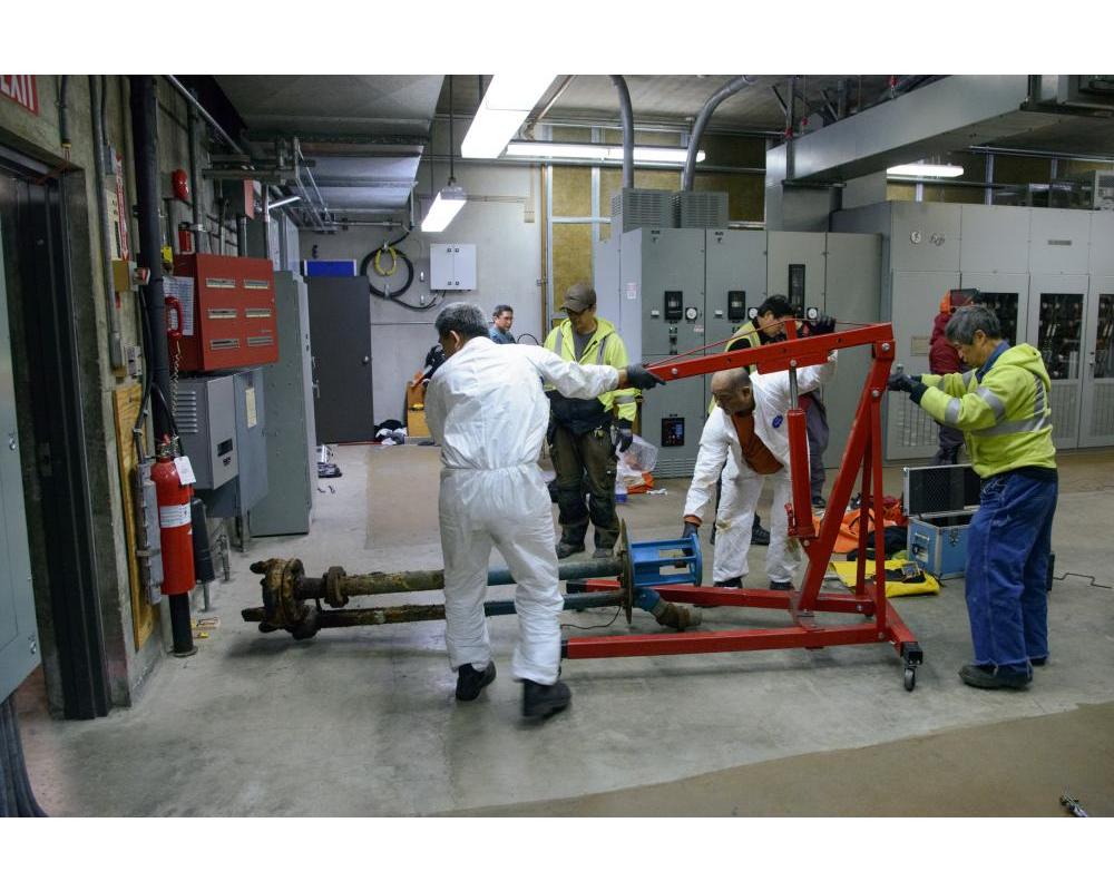 Several crew members holding a large device instrument inside a systems shop location. 