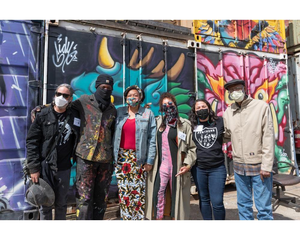 Several individuals with masks huddle for a group photo in front of a colorful mural backdrop in an our setting. 