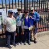 a group of people stand before a tall iron fence in front of informational boards. They smile at the camera. 