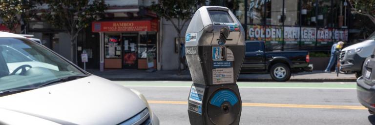 Image shows a parking meter on a San Francisco street. A parked car is in the foreground, with shops in the background.