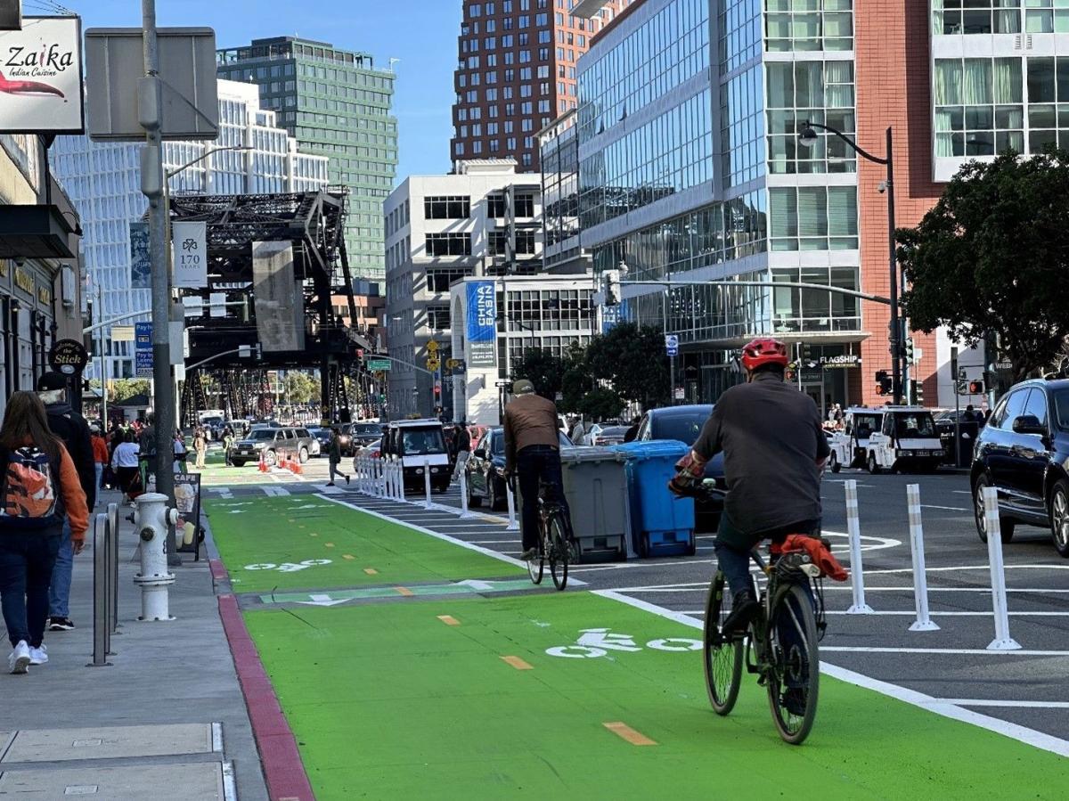 People walking on the sidewalk and riding bikes on a two-way bike lane towards a bridge on a busy street.