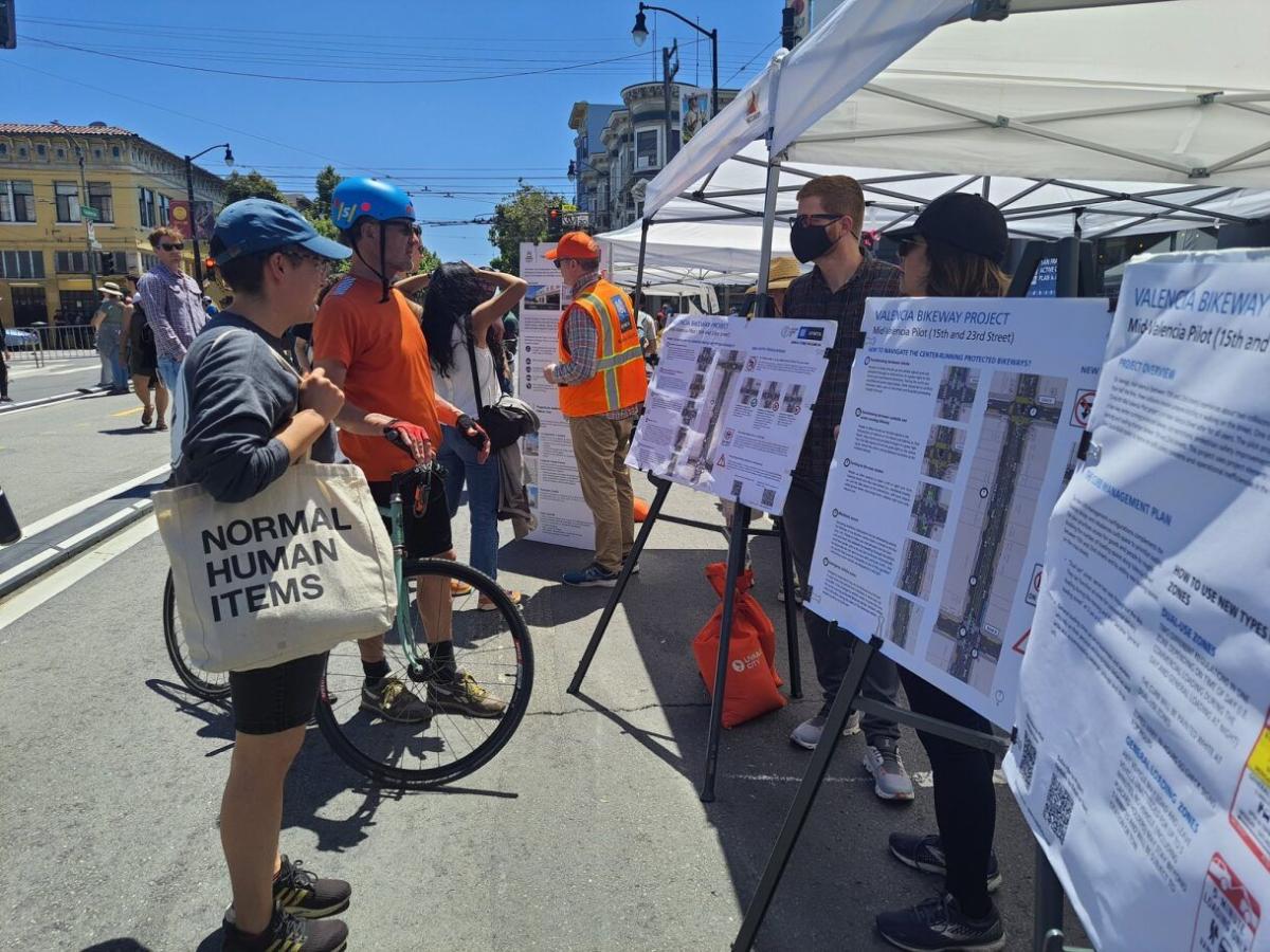 Outreach staff talk to community members in front of signs for the Valencia center-running bikeway