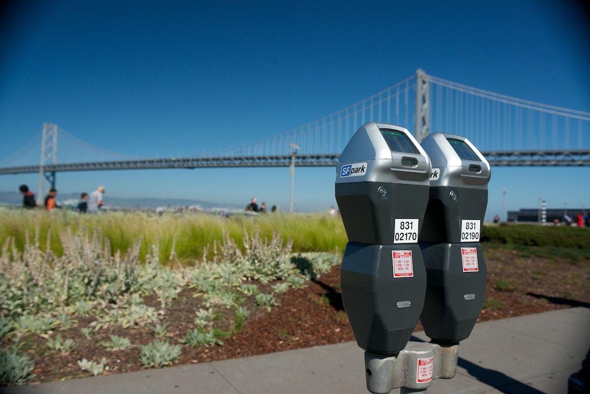 Close-up of two parking meters in front of a grassy area and the Bay Bridge in the background.