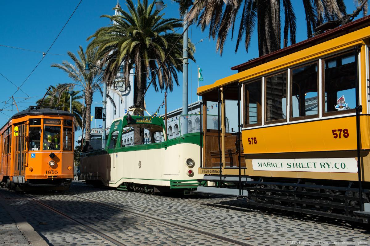 An orange historic streetcar passes a green and cream open-air Boat Tram and another orange historic streetcar. Palm trees and Ferry Building in the background.