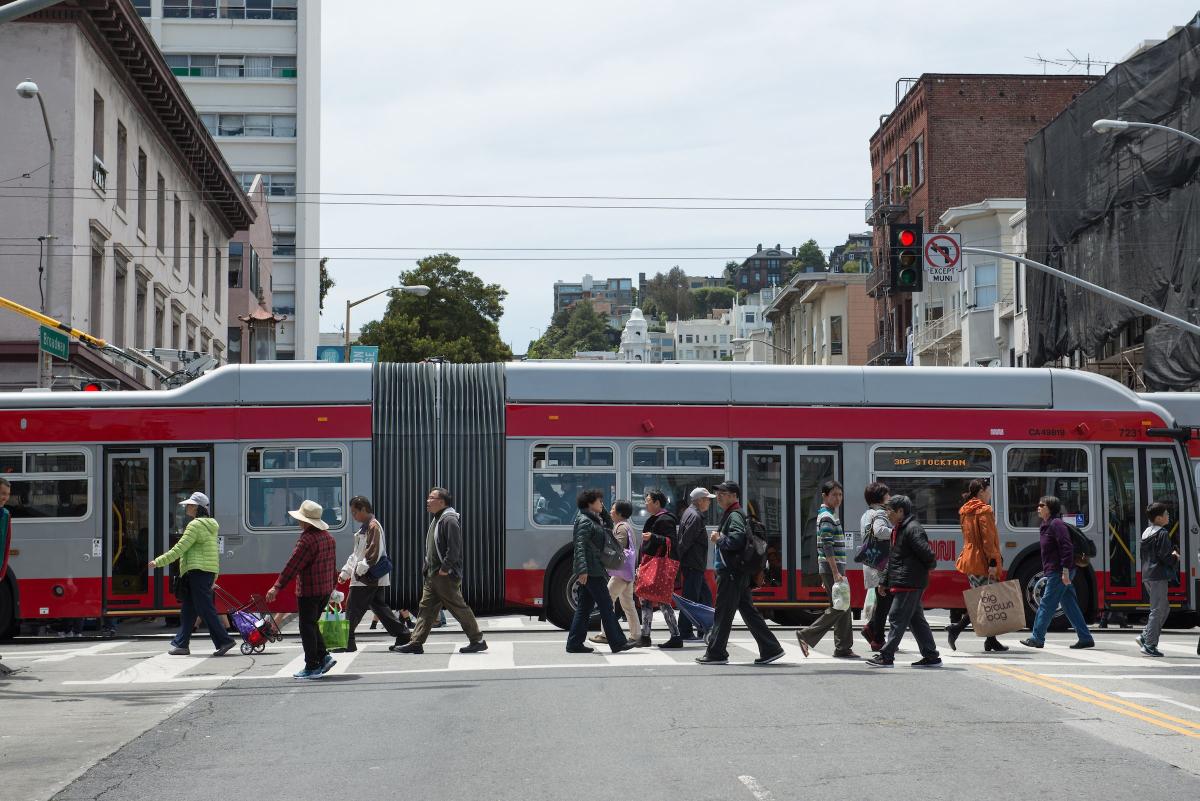 People cross the street as a bus passes them in the background in the same intersection.