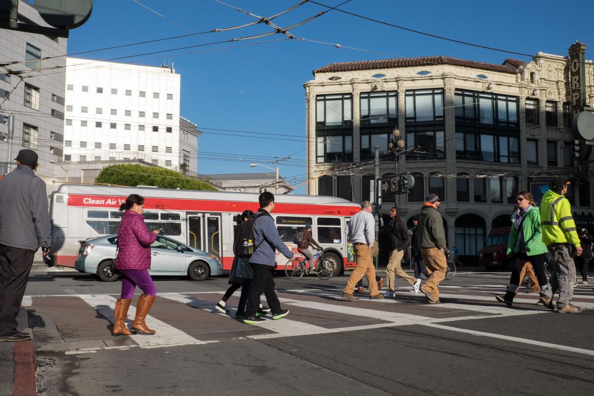 People cross a street as cars and buses and people riding bikes travel in the lanes near them.