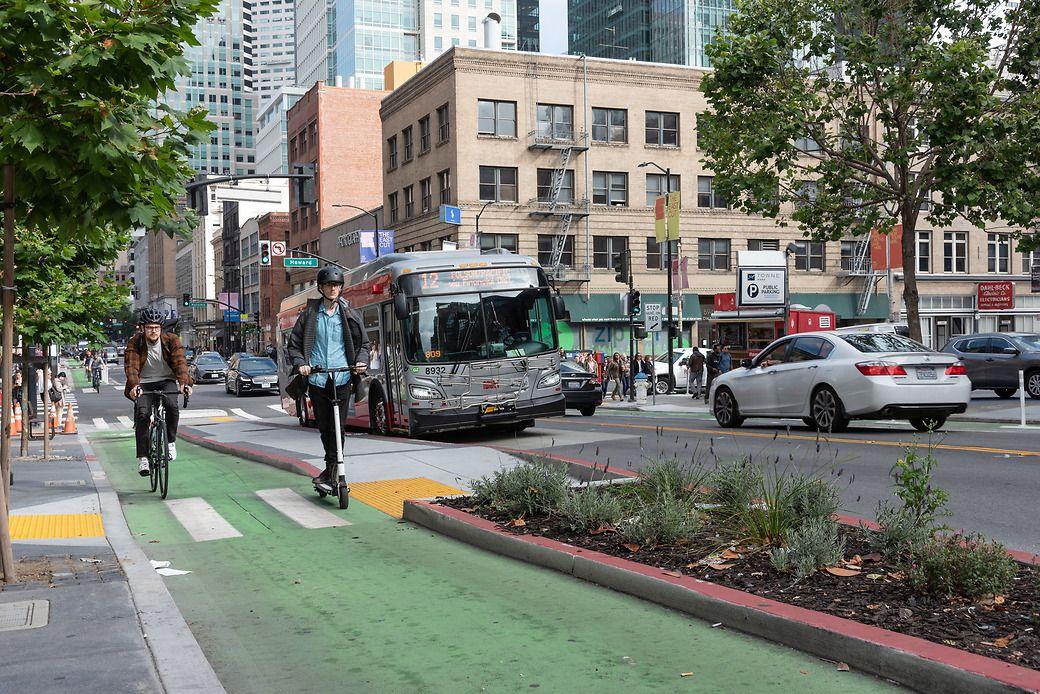 Scooter riders use a bikeway that's painted green on a city street. Buses and cars are in the background.