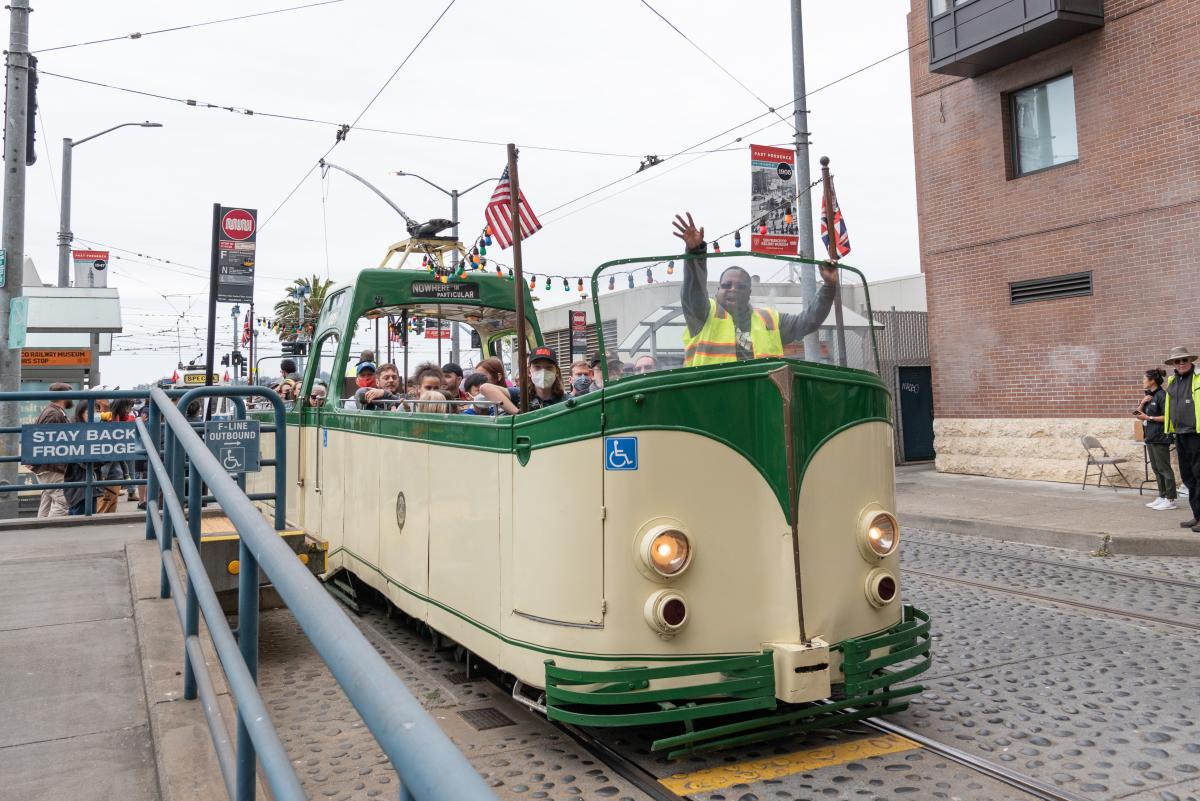 Cream and green open-air streetcar, a Boat Tram, drives near the Embarcadero. The operator waves.