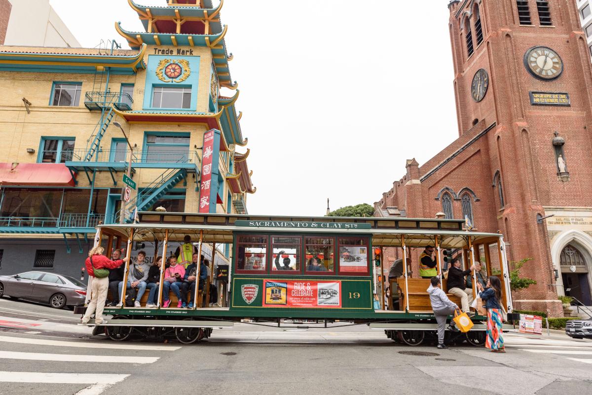 Cable car with a sign that says "Sacramento & Clay St" stops in Chinatown near a red brick church. 