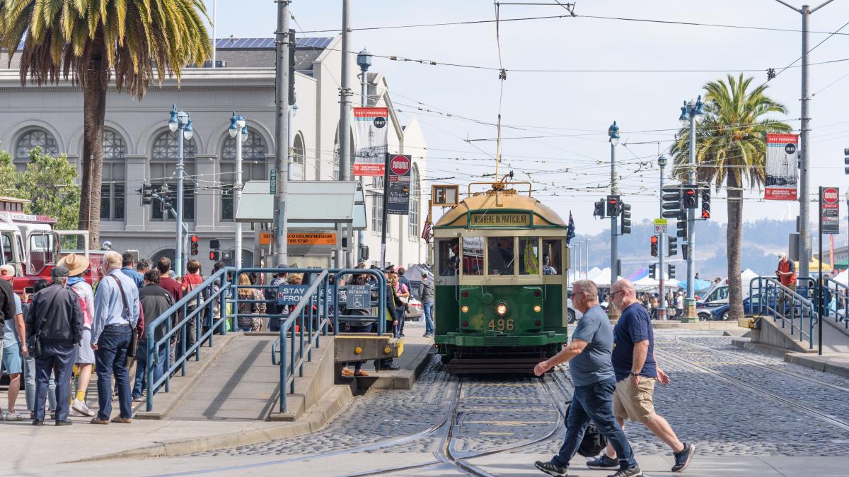 Green and cream historic streetcar stops near the Embarcadero. People cross an intersection in front of the streetcar.