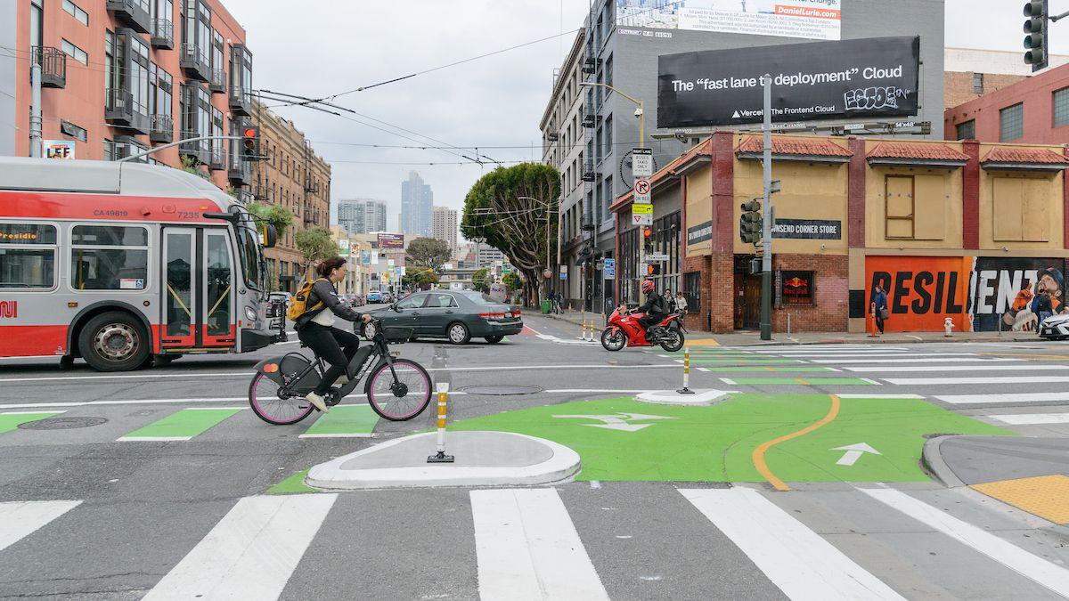 Person rides bike on a bikeway that' painted green in SoMa. In the background, a bus, vehicle and motorcycle cross the same intersection.