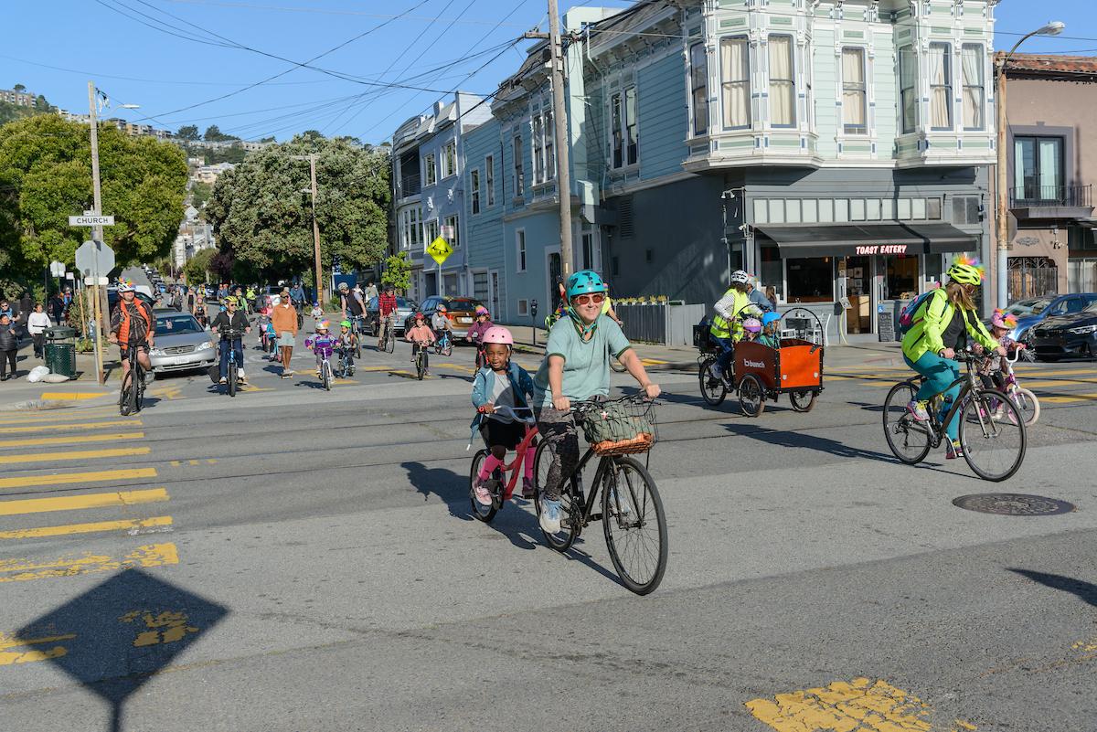 Adults and students ride their bikes, crossing a street on a sunny day.