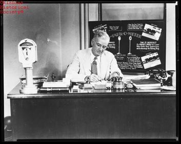 Photo of Carl C. Magee, the inventor of the parking meter, sitting at his work desk. A meter prototype rests on the desk.