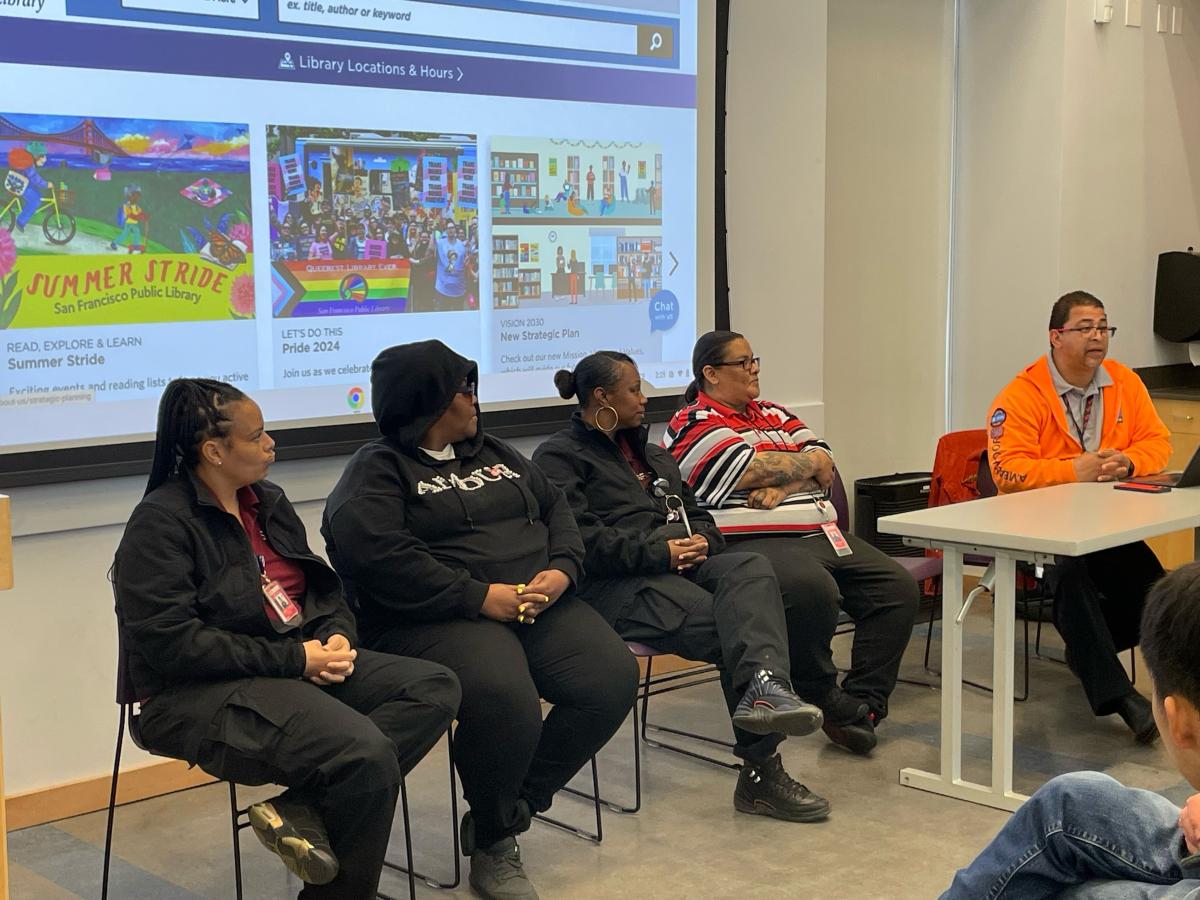 Muni staff sit in chairs during a panel talk with a projector behind them showing a presentation.