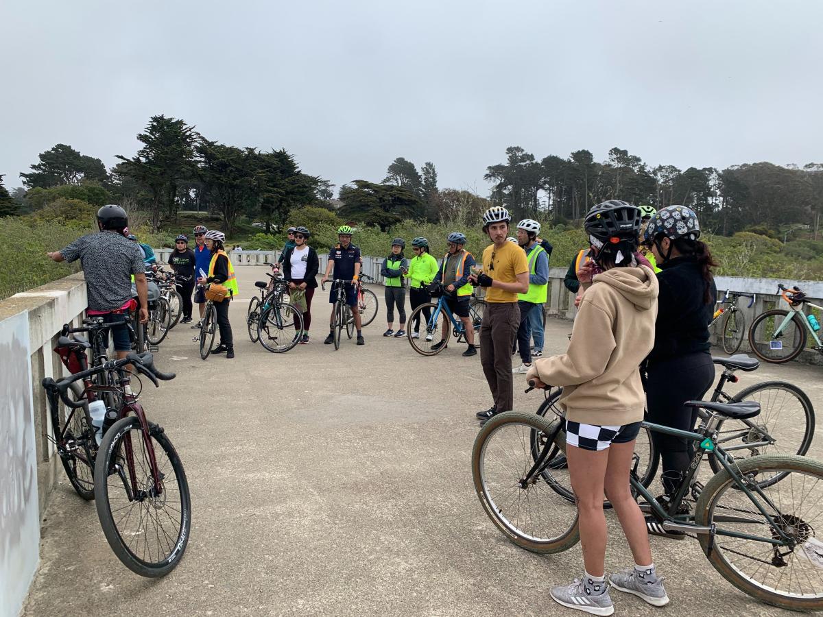 People gather outside, standing by their bikes. We see lots of trees and grass in the background.