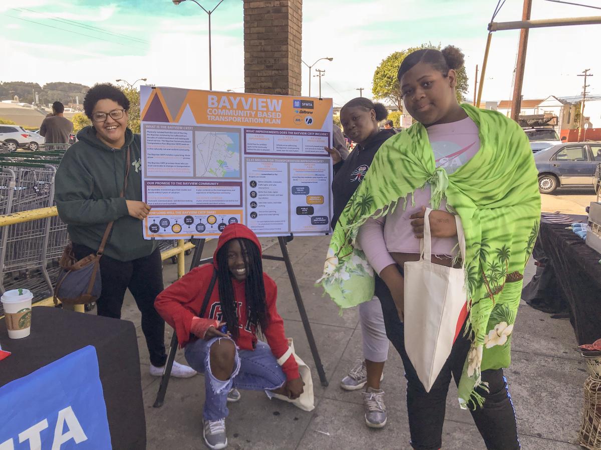 Students smile around a sign featuring information about the Bayview Community Based Transportation Plan. 