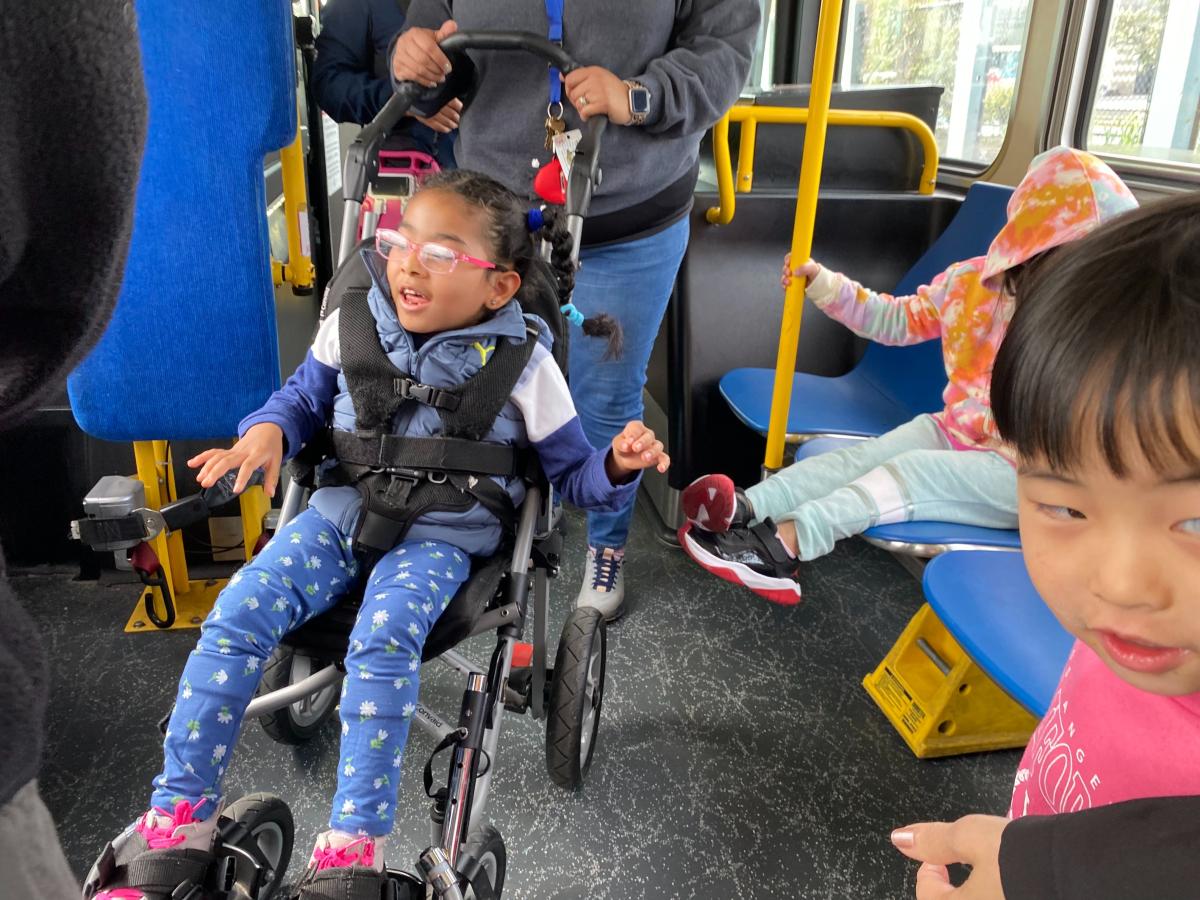 A passenger in a mobility stroller smiles near other young children on a bus.