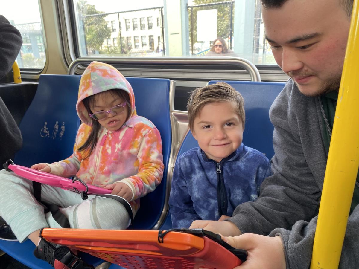 Two young children on a bus pay attention to an adult who shows them route information on a phone.