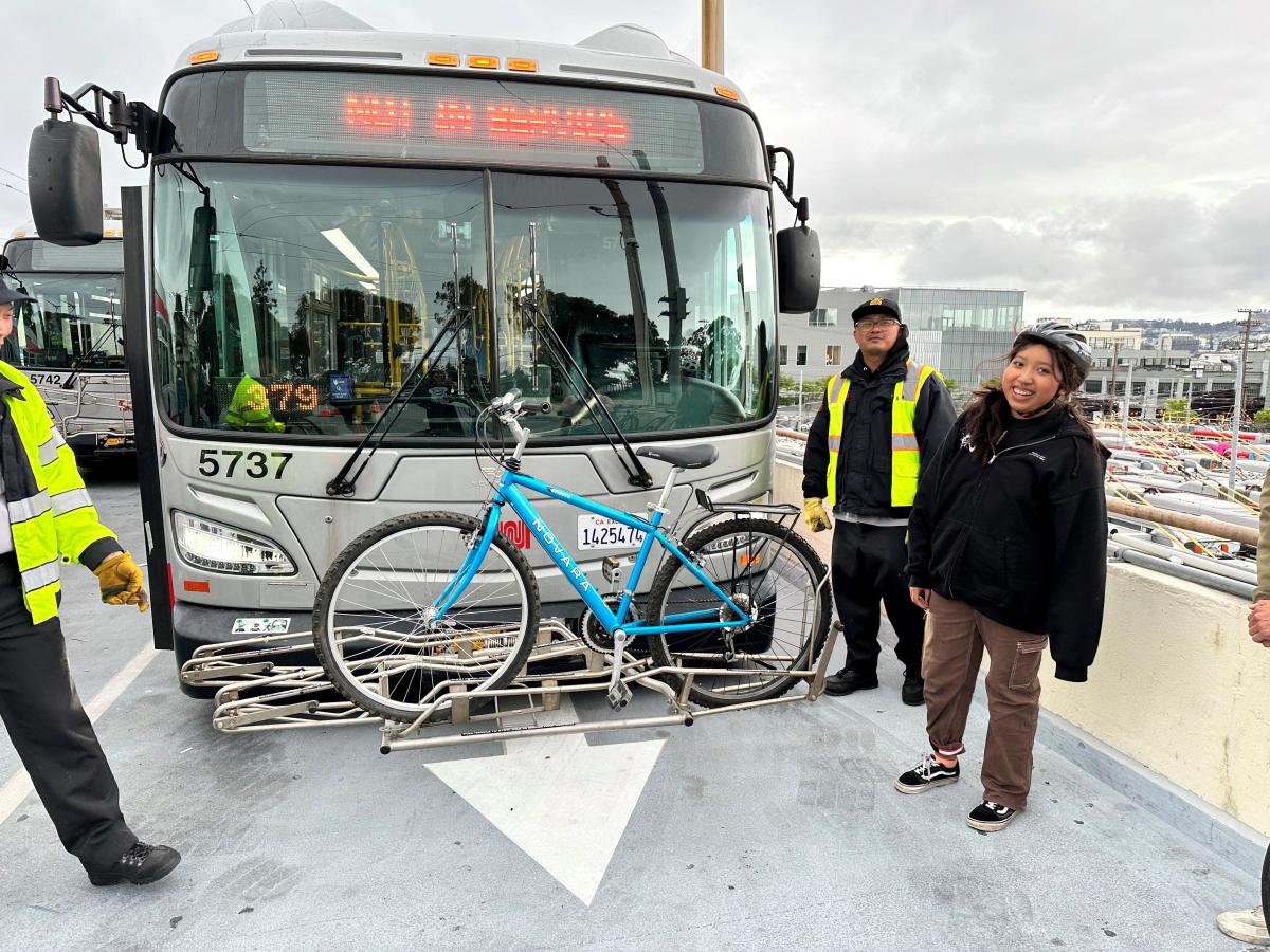 Muni bus has bike stored on the front rack. A rider with a helmet smiles near two Muni staff, who stand by the bus.