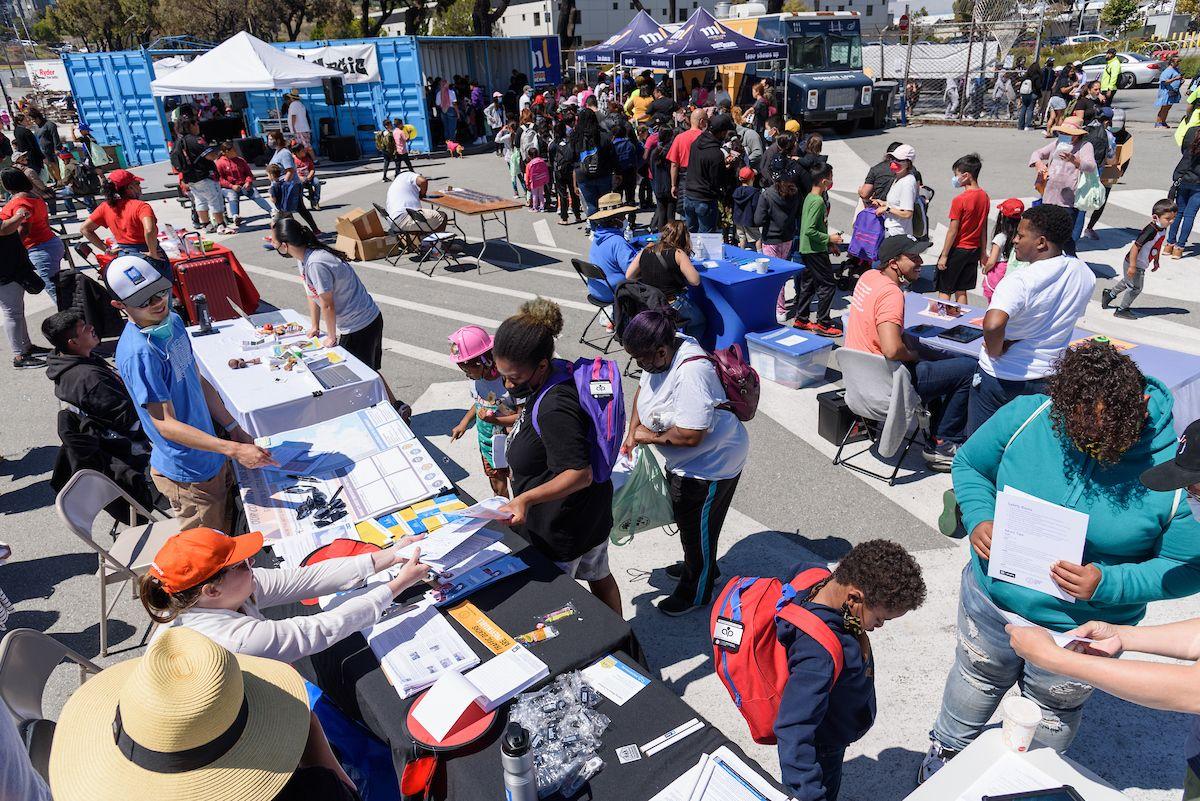 Overhead view of an outdoor public event where SFMTA staff and community members chat over tables about the Bayview Community Shuttle.
