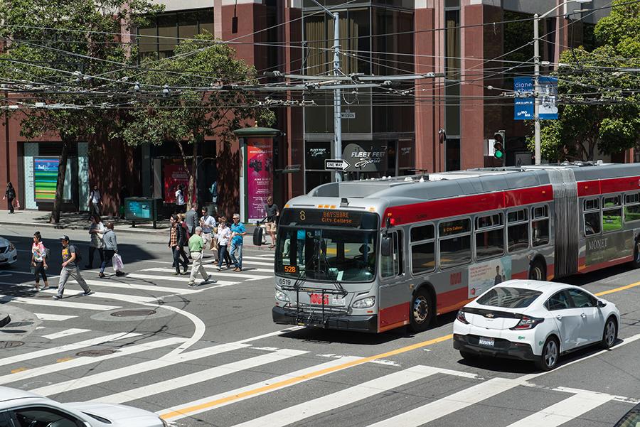 8 San Bruno bus crosses an intersection alongside people walking in the crosswalk.