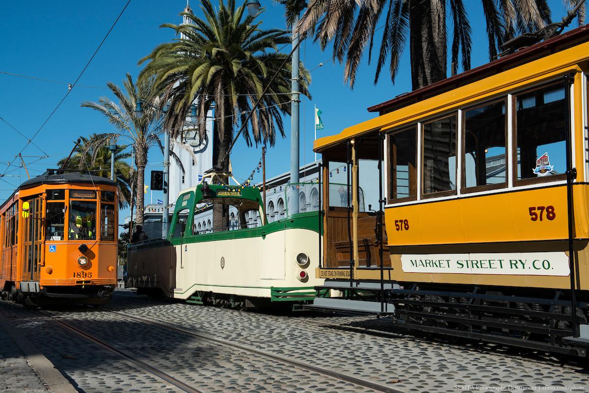 Three retro streetcars, including the Boat Tram, stop near palm trees by the Ferry Building.