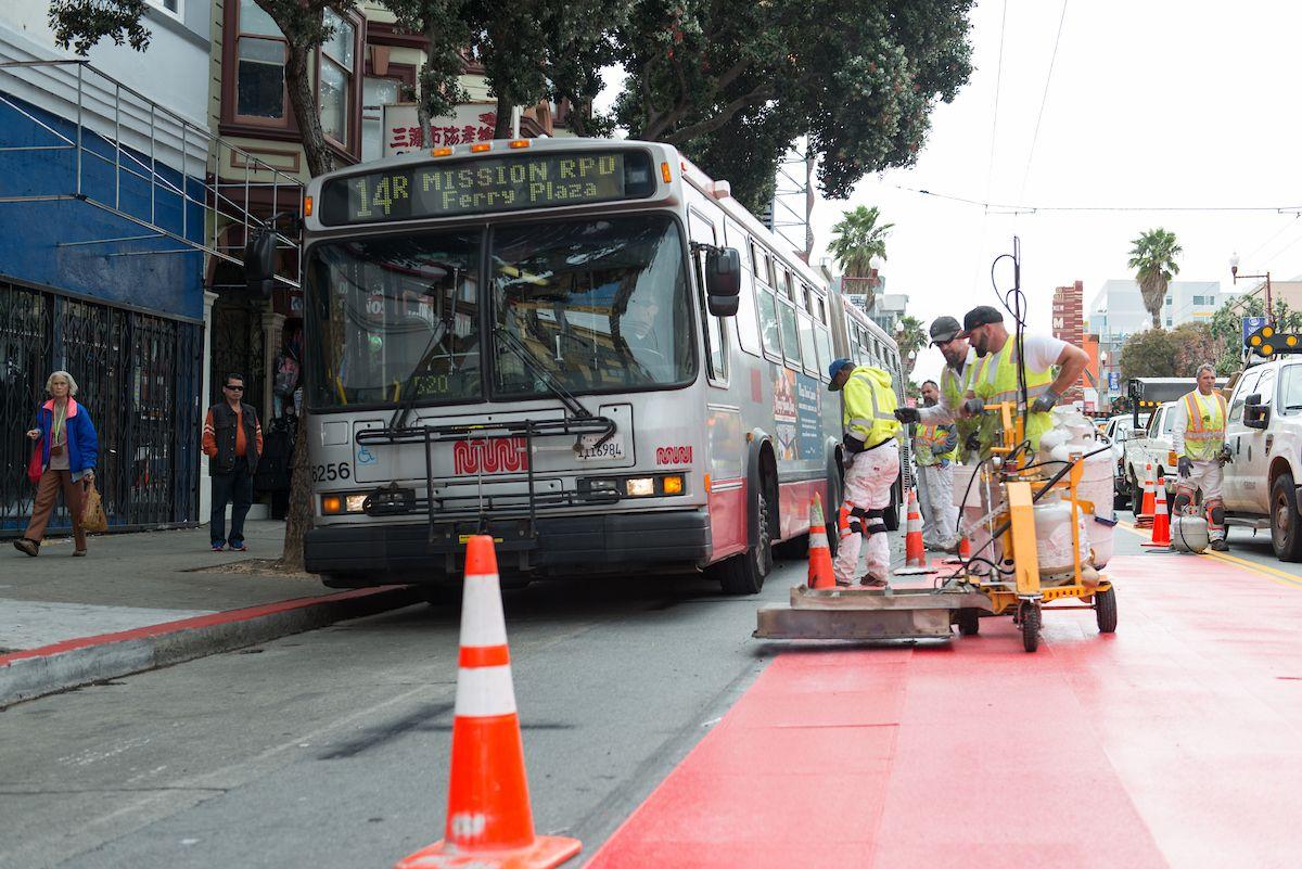 A photograph of an SFMTA painting crew installing a red transit lane on Mission Street. The 14R Mission Rapid can be seen to the left of the paint crews. 