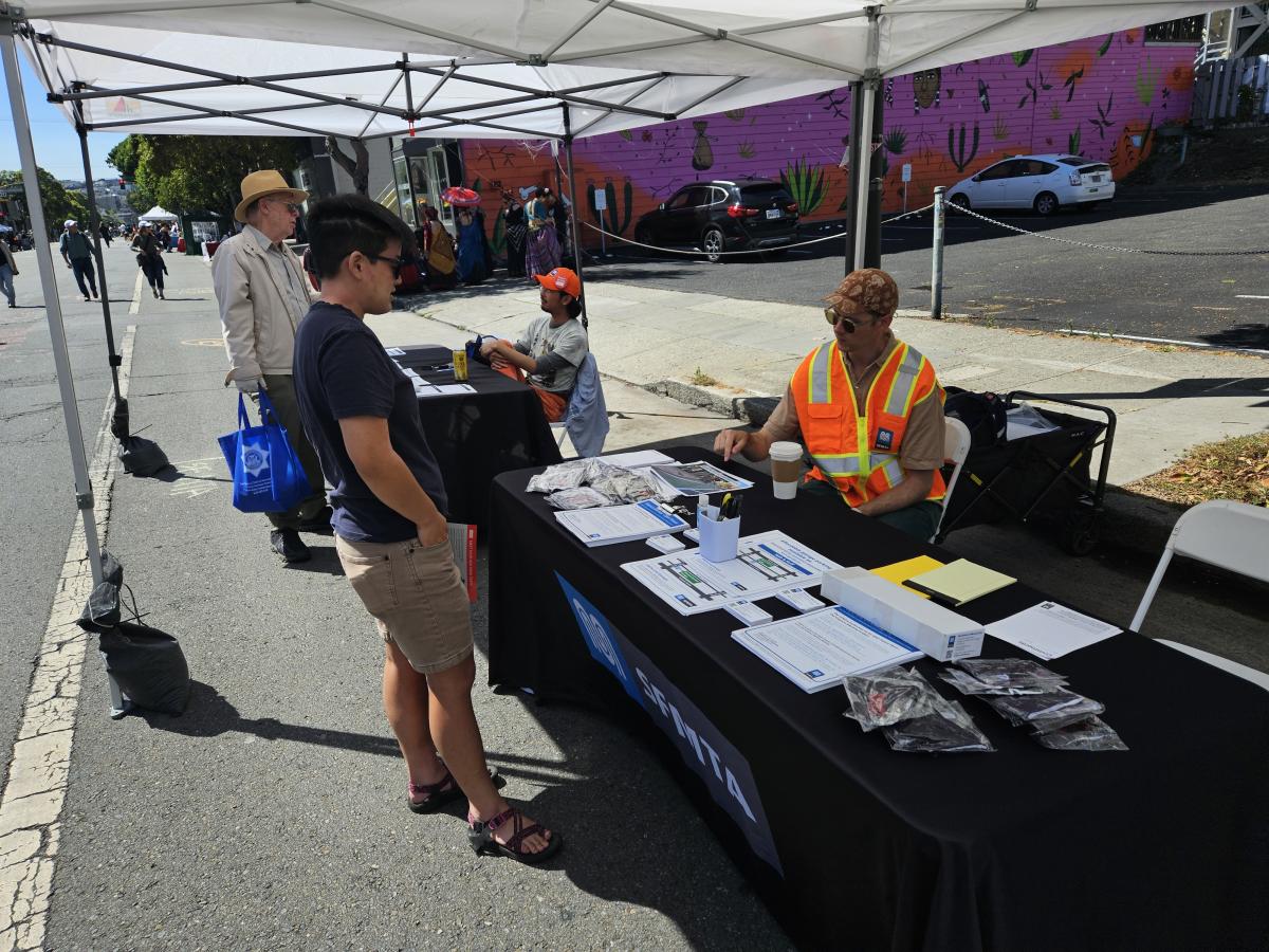 A member of the public is shown talking to an SFMTA staff person wearing an orange safety vest. A table, between them, is covered with diagrams and papers about the project.