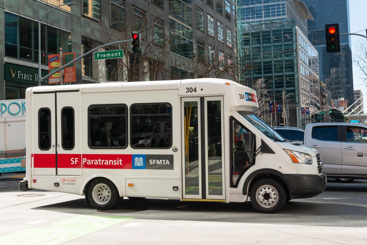 A SF Paratransit van crosses a downtown intersection along with other vehicles.