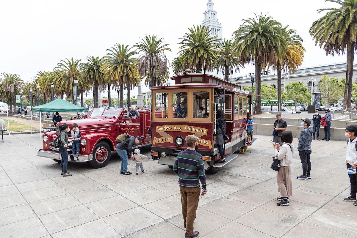 Several people stand near two retro vehicles. One is a cable car and the other is a red, open air vehicle.