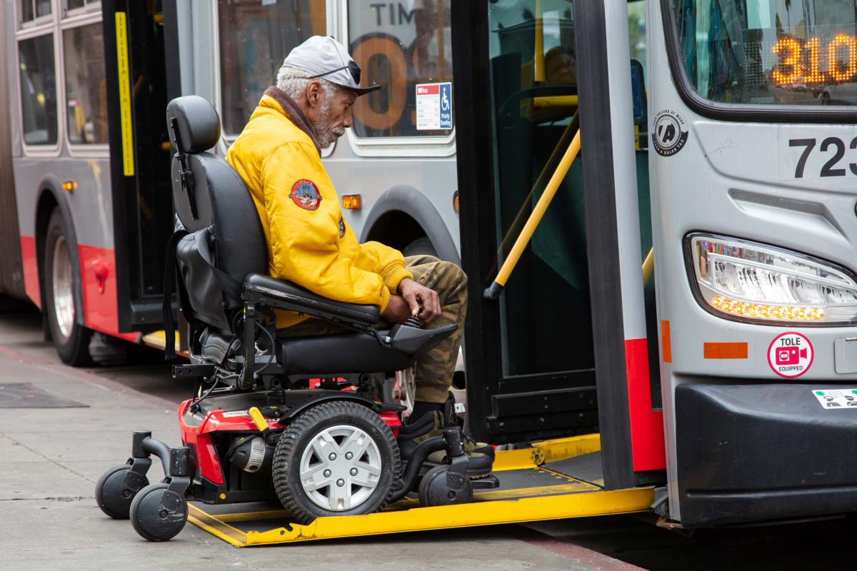 Person using a mobility device boards a Muni bus through a ramp lowered to sidewalk level.