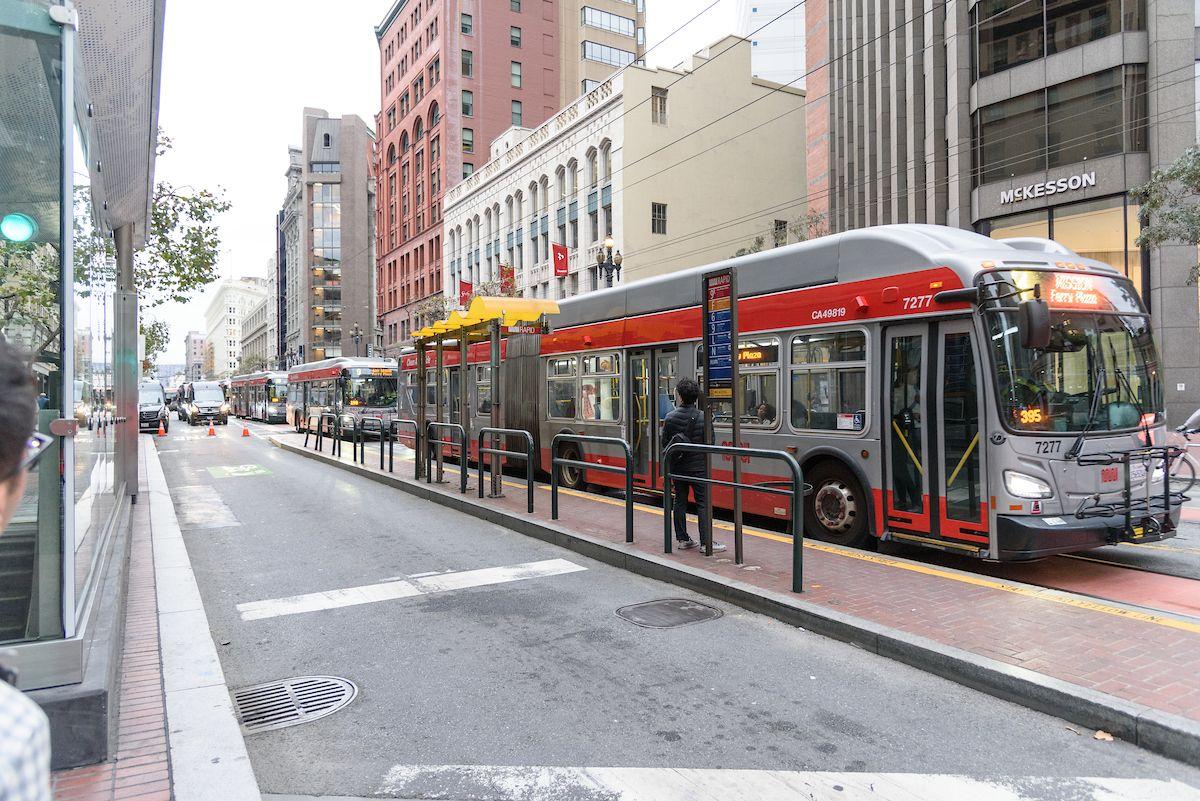 A photograph of buses lined up on Market Street near a boarding platform. A person is waiting on the platform. Boarding platforms help people get on and off the bus quickly and safely. 