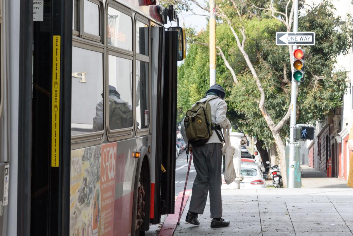 Person wearing a backpack and using a mobility device exits a Muni bus. 