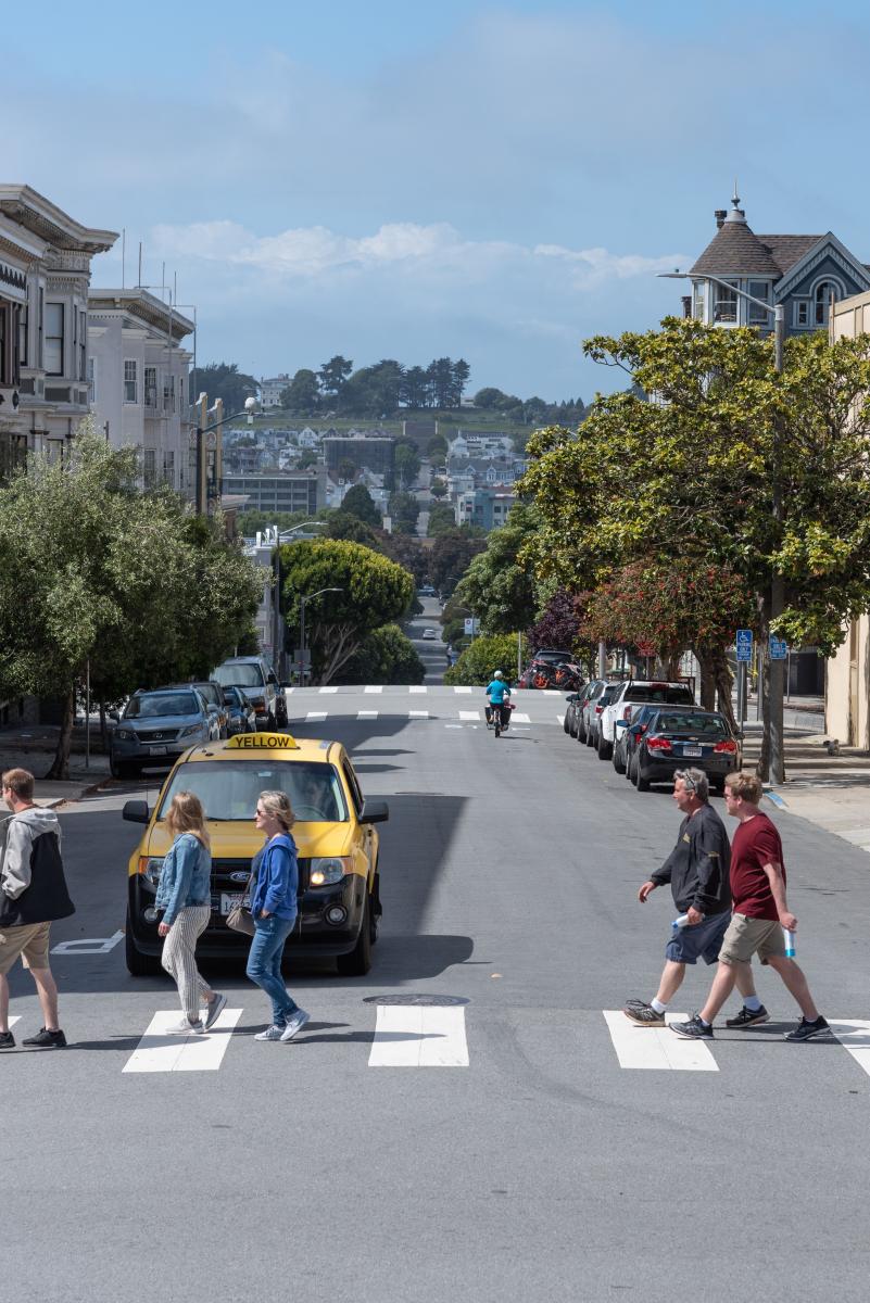 Several people cross an intersection as a taxi driver waits behind the crosswalk. Alamo Square is seen in the background.