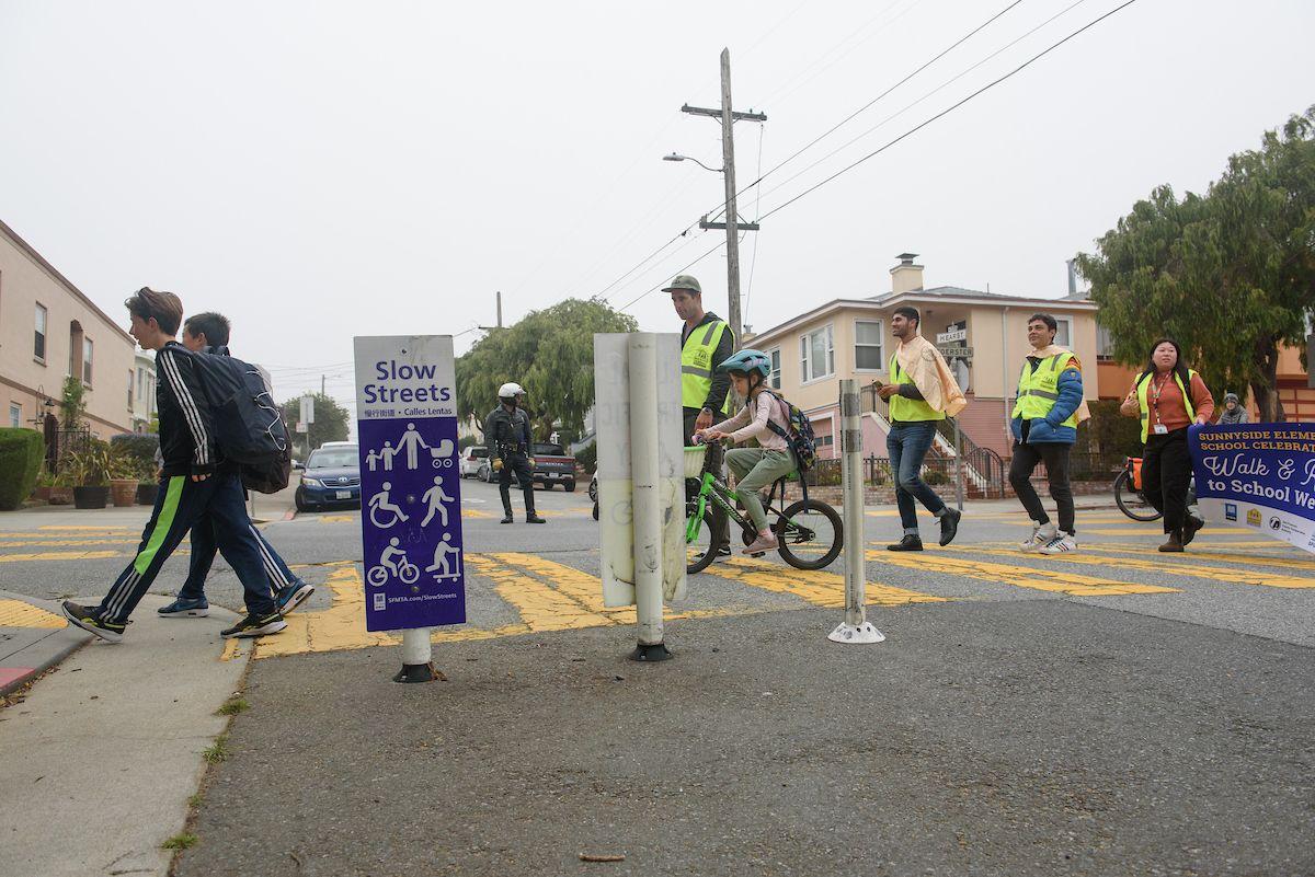 Students walk and ride bikes to cross a Slow Street alongside adults wearing yellow safety vests. Students walk and ride bikes to cross a Slow Street alongside adults wearing yellow safety vests.